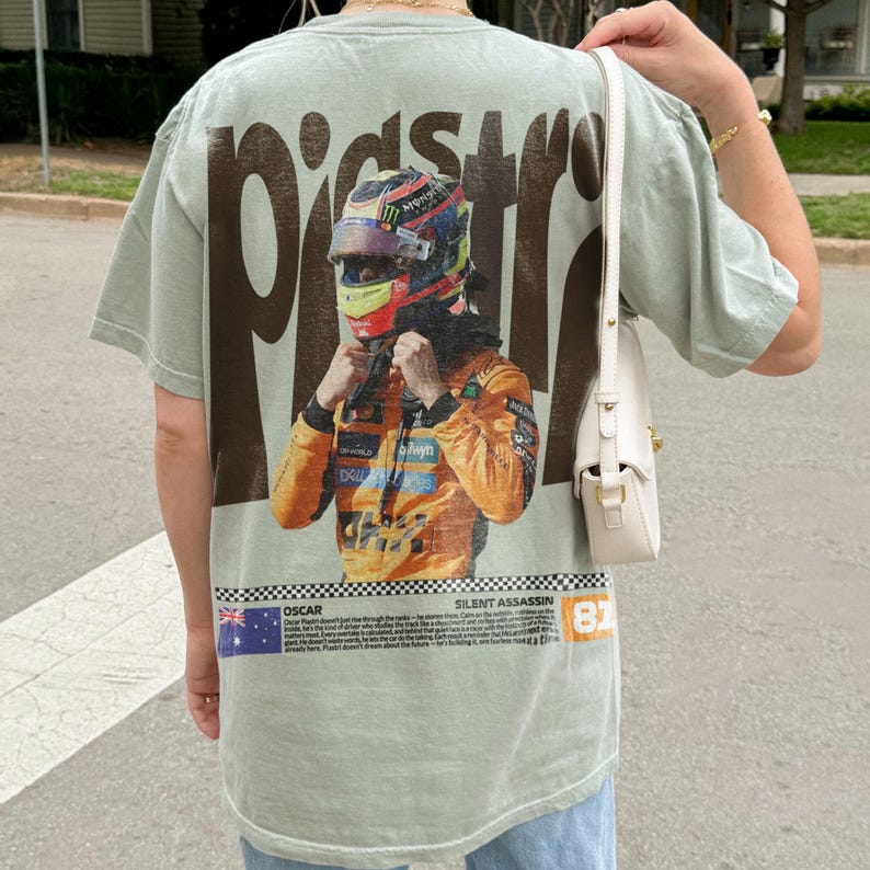 May include: Sage green t-shirt featuring a graphic of a race car driver in a helmet and racing suit. The word "PISTONS" is printed in large brown letters above the graphic. The bottom of the shirt has text and the number 81.