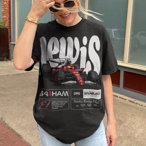 Pode incluir: T-shirt preto com um design gr&aacute;fico de um carro de corrida de F&oacute;rmula 1 em vermelho e preto. A camisa tem o nome "LEWIS" em letras brancas grandes e inclui o texto "44 HAM DRS ENABLED" e "Sunday Racing Club".