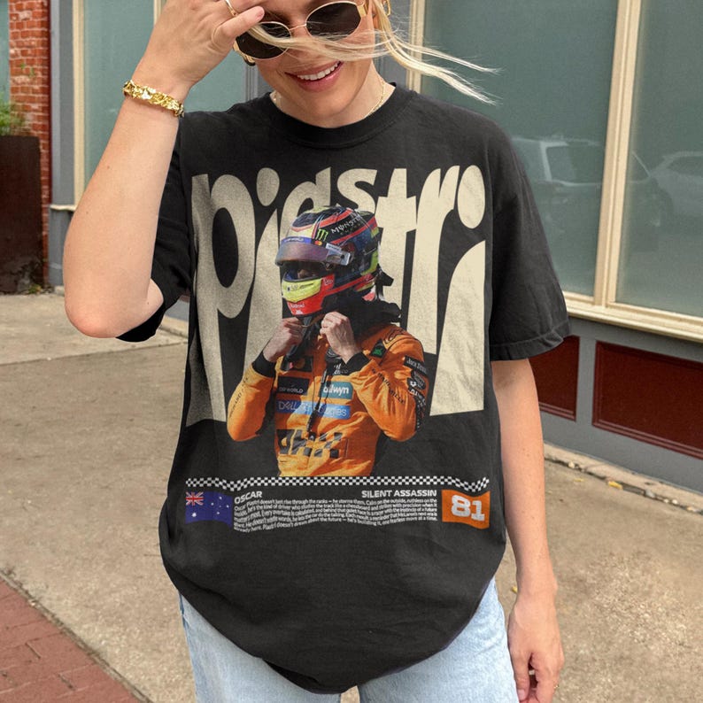 May include: Black graphic t-shirt featuring a Formula 1 driver in an orange racing suit and helmet. The shirt has the text "Oscar" and "Silent Assassin" with the number "81". The shirt is worn by a person.