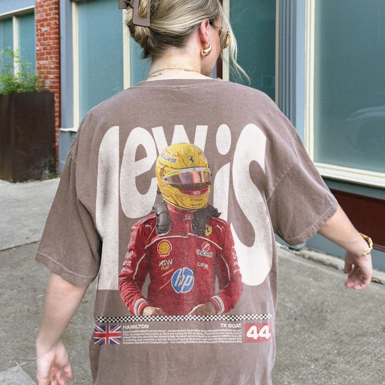 May include: Brown t-shirt with a graphic of a Formula 1 driver in a red racing suit and yellow helmet. The word "LEWIS" is printed in large white letters across the back. The shirt also includes the Union Jack flag and the number 44.