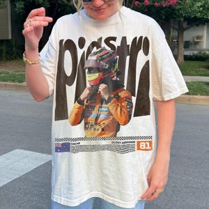 May include: Off-white t-shirt with a graphic of a race car driver in a helmet and orange racing suit. The word "PISSTR" is printed in large, dark brown letters above the image. Below the image is the text "OSCAR SILENT ASSASSIN 81".
