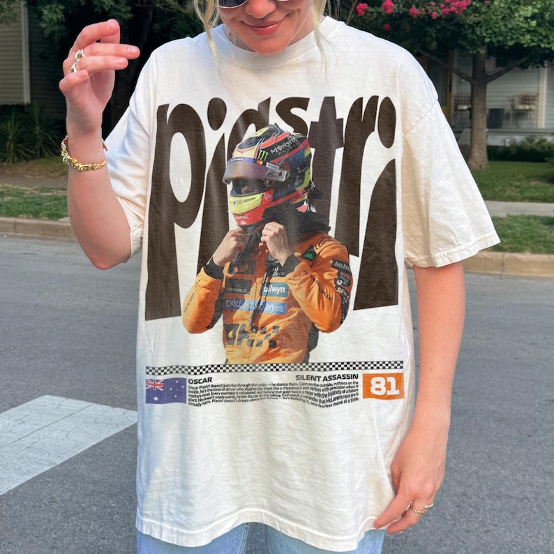 May include: Off-white t-shirt with a graphic of a race car driver in a helmet and orange racing suit. The word "PISSTR" is printed in large, dark brown letters above the image. Below the image is the text "OSCAR SILENT ASSASSIN 81".