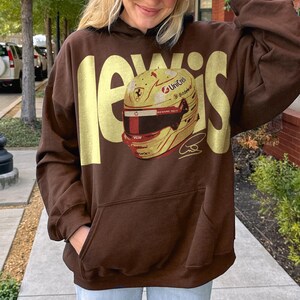 May include: Brown hoodie with the name "Lewis" in large, gold letters. The hoodie features a graphic of a racing helmet with sponsor logos and a signature. The hoodie has a front pocket and a hood.