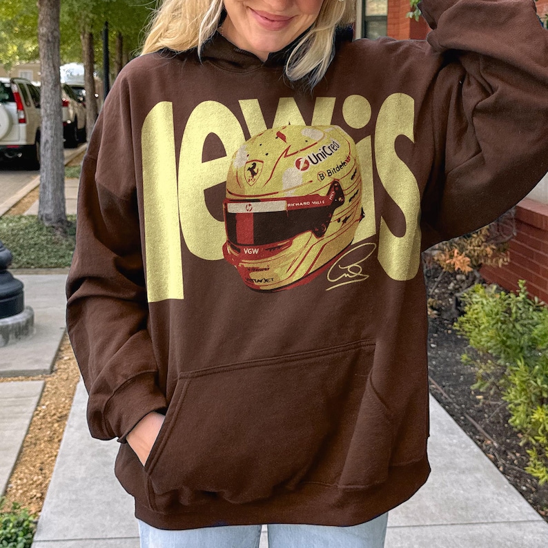 May include: Brown hoodie with the name "Lewis" in large, gold letters. The hoodie features a graphic of a racing helmet with sponsor logos and a signature. The hoodie has a front pocket and a hood.