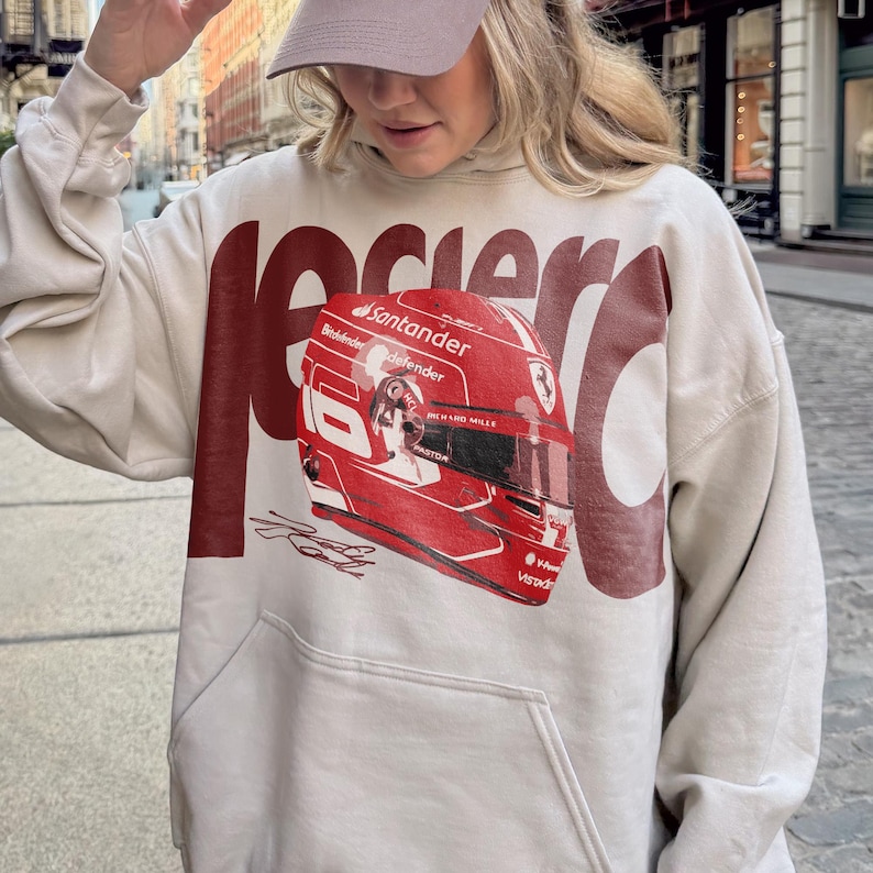 May include: Beige hoodie with a large red graphic of a racing helmet. The helmet has the Santander logo and the word "defender" printed on it. The word "Racer" is printed in large red letters above the helmet.