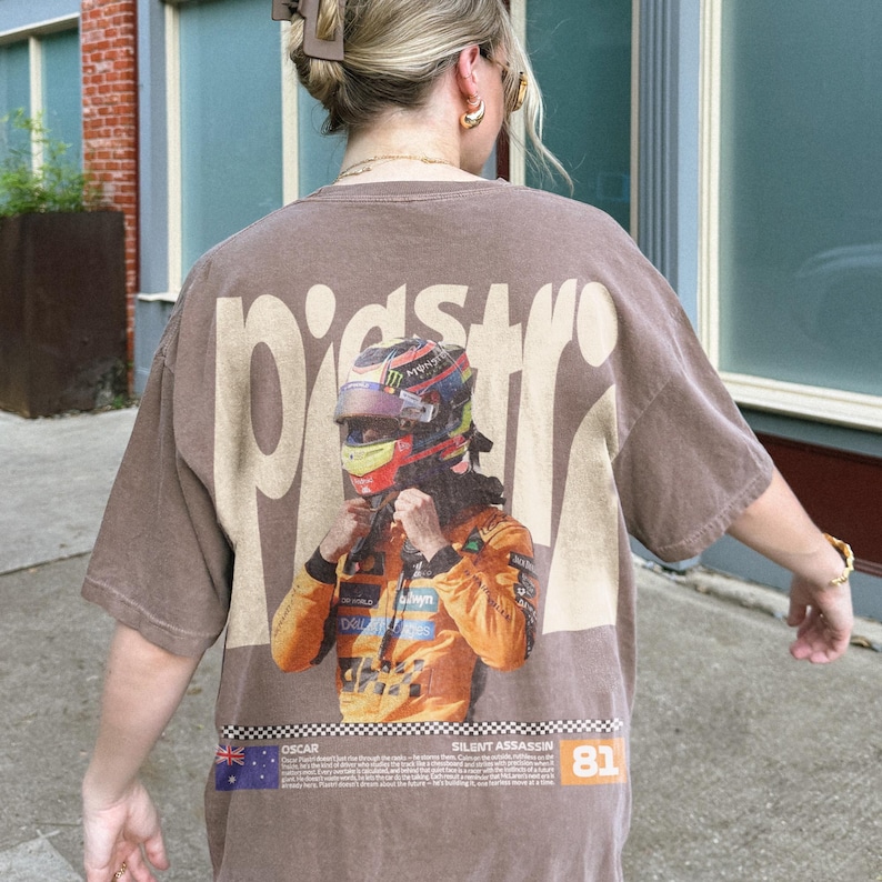 May include: Brown t-shirt with a large graphic on the back featuring a race car driver in a helmet and racing suit. The word "PIST" is printed above the graphic. Below the graphic is the text "OSCAR" and "SILENT ASSASSIN" with the number "81".