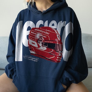 May include: Navy blue hoodie featuring a red and white racing helmet graphic. The helmet has sponsor logos and the number 16. The word "Racer" is printed in large white letters across the top.