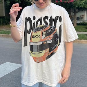 May include: Cream-colored t-shirt with the word "pidstr" in large black letters. The shirt features a graphic of a racing helmet in yellow, orange, black, and white. The helmet has the words "Monster" and "McLaren" printed on it.