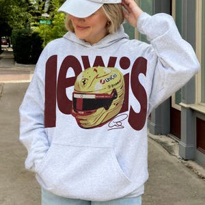 May include: A light gray hoodie with a large maroon "Lewis" graphic. The graphic features a yellow and gold racing helmet with sponsor logos and a signature. The hoodie has a front pocket and a drawstring hood.