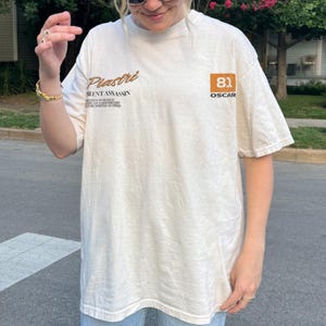 May include: Cream-colored t-shirt with the text "Piastri SILENT ASSASSIN" in brown script. The shirt also features a brown square with the number "81" and the word "OSCAR". The shirt is a casual, oversized fit.