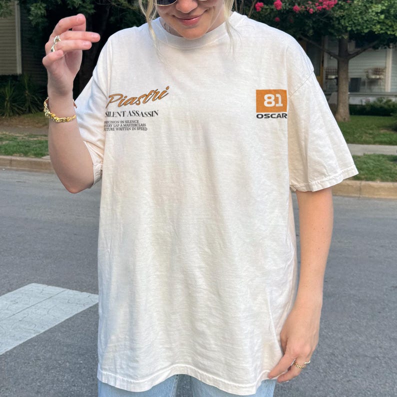 May include: Cream-colored t-shirt with the text "Piastri SILENT ASSASSIN" in brown script. The shirt also features a brown square with the number "81" and the word "OSCAR". The shirt is a casual, oversized fit.