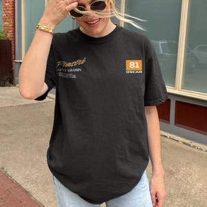 May include: Black t-shirt with the text "Piastri Silent Assassin" in gold script on the upper left chest. The right chest features a square orange box with the number "81" and the word "OSCAR" below it. The shirt is a crew neck and has short sleeves.