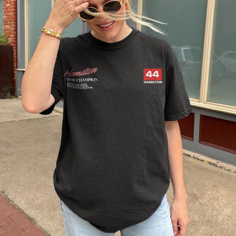 May include: Black t-shirt with the name "Hamilton" in red script and the number "44" in a red box. The shirt also has text that reads "TIME CHAMPION". The short-sleeved shirt is a casual style.