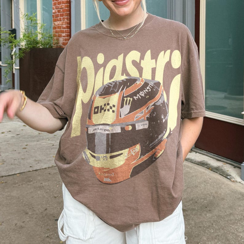 May include: Brown t-shirt with the word "Pidstro" in gold lettering. The shirt features a graphic of a racing helmet with orange and black accents. The person is wearing white shorts.