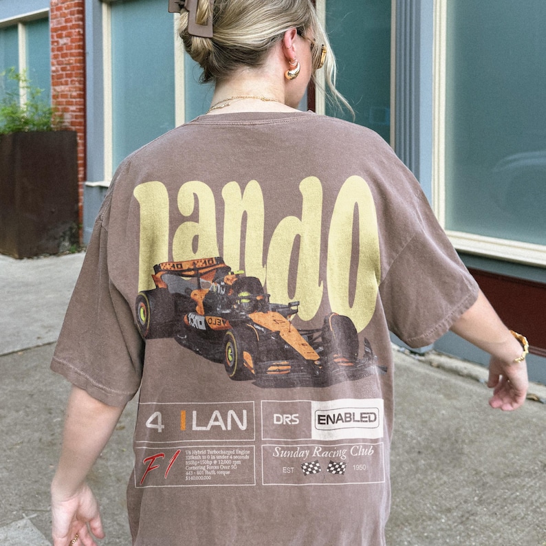 Pu&ograve; includere: T-shirt marrone con una grande grafica "Lando" gialla sul retro. Sotto, una grafica di una macchina da corsa di Formula 1 nera e arancione. Il testo aggiuntivo include "4 LAN DRS ENABLED" e "Sunday Racing Club EST 1950".