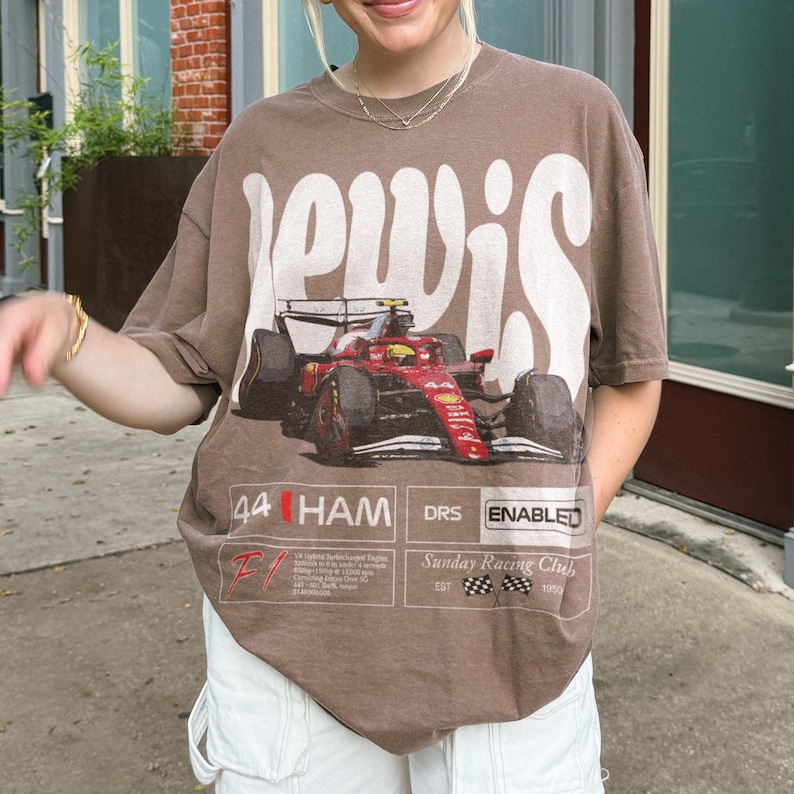 Pode incluir: Camiseta marrom com a palavra "Lewis" em letras brancas grandes. A camiseta apresenta um gr&aacute;fico de um carro de corrida vermelho e o texto "44 HAM DRS ENABLED" e "Sunday Racing Club".