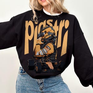 K&ouml;nnte beinhalten: Schwarzes Sweatshirt mit einem goldenen Aufdruck eines Rennfahrers in Helm und Rennanzug sowie einem Rennwagen. Das Wort "Piastri" ist in gro&szlig;en goldenen Buchstaben gedruckt. Das Sweatshirt wird von einer Person getragen.