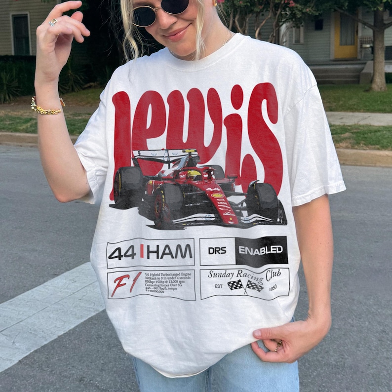 Pode incluir: Camiseta branca com um gr&aacute;fico vermelho de um carro de corrida de F&oacute;rmula 1 e o nome "Lewis". A camisa tamb&eacute;m apresenta o texto "44 HAM DRS ENABLED" e "Sunday Racing Club", com um design de bandeira quadriculada.