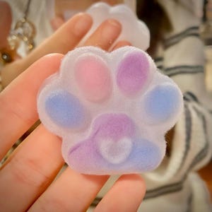 Pode incluir: Um acessório em forma de pata de gato em tons pastel, com almofadas rosa, roxas e azuis e um detalhe de coração. O acessório é segurado em uma mão, com uma textura macia e difusa.