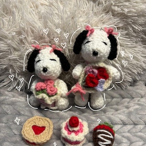 Op de afbeelding: Gehaakte Snoopy-poppen met zwarte oren en roze strikken, met boeketten en omringd door gehaakte desserts: een hartvormige koek, een cake en een chocolade-aardbei. De poppen en desserts zijn wit, roze, rood en bruin.