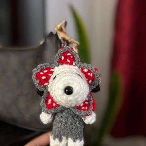 Op de afbeelding: Een handgemaakte gehaakte Demogorgon sleutelhanger. De Demogorgon heeft een witte kop, een grijze body en rood-witte bloembladachtige kenmerken. Het heeft een zwarte knoopneus en kleine zwarte accenten. De sleutelhanger is bevestigd aan een goudkleurige ketting.