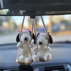 May include: Two white crochet dog ornaments with black ears and eyes hang from a car's rearview mirror. Each dog is seated on a small swing made of brown yarn. One dog has a pink bow, the other a purple bow.