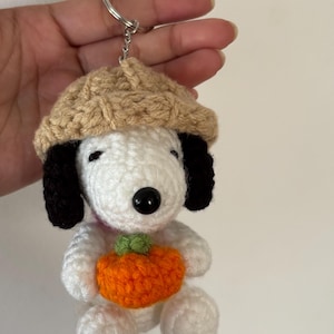 Op de afbeelding: Gehaakte Snoopy sleutelhanger met een bruine hoed en een oranje pompoen. De wit en zwarte Snoopy figuur heeft een zwarte knoopneus en zwarte oren. De pompoen heeft een groene steel. De sleutelhanger is bevestigd aan een zilveren ring.