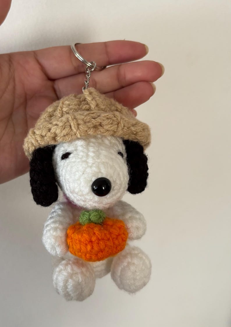 Op de afbeelding: Gehaakte Snoopy sleutelhanger met een bruine hoed en een oranje pompoen. De wit en zwarte Snoopy figuur heeft een zwarte knoopneus en zwarte oren. De pompoen heeft een groene steel. De sleutelhanger is bevestigd aan een zilveren ring.