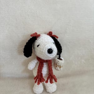 Op de afbeelding: Gehaakte Snoopy-figuur met zwarte oren, een rode sjaal en twee rode strikken. De witte hond houdt een klein, beige kopje vast. Het figuur is handgemaakt en heeft een schattig, grillig ontwerp.