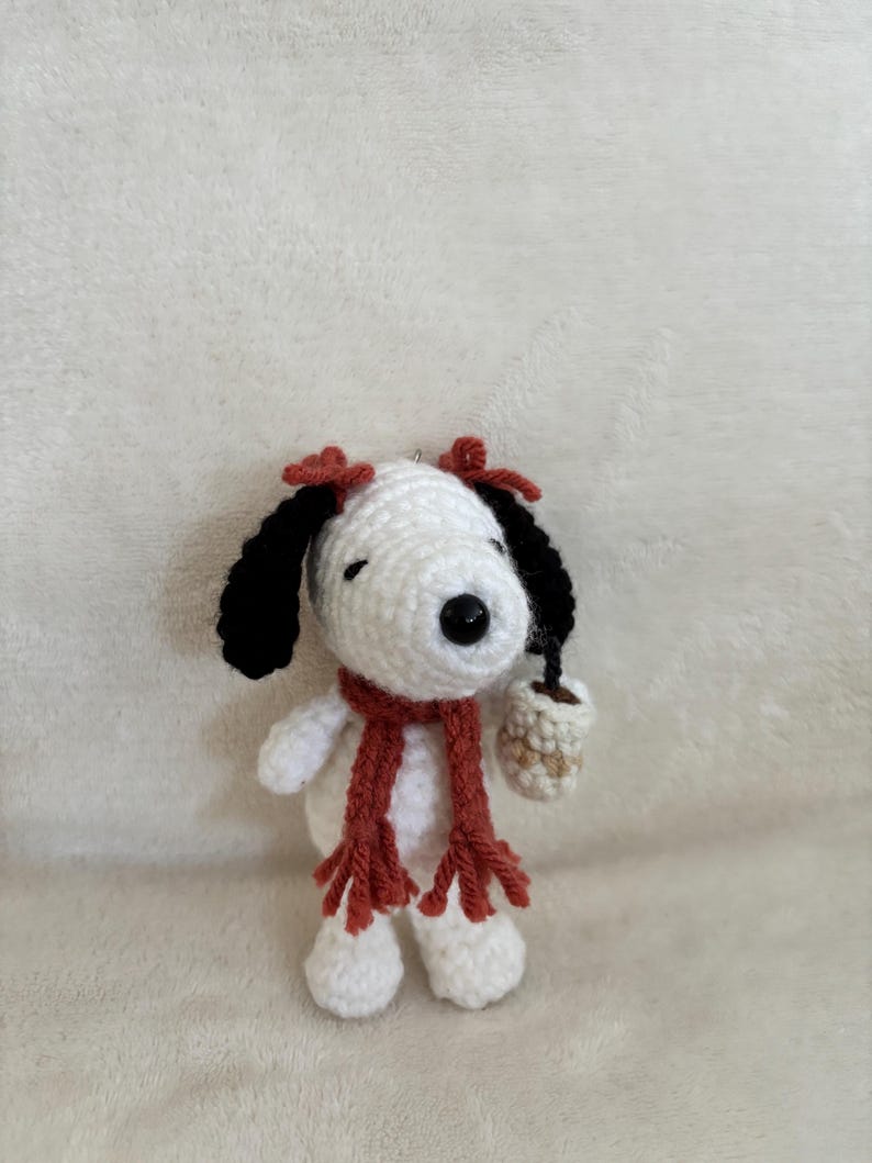 Op de afbeelding: Gehaakte Snoopy-figuur met zwarte oren, een rode sjaal en twee rode strikken. De witte hond houdt een klein, beige kopje vast. Het figuur is handgemaakt en heeft een schattig, grillig ontwerp.