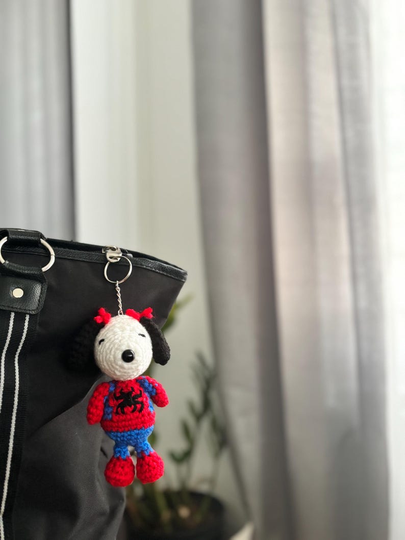 May include: A handmade crochet keychain featuring a cartoon dog character in a Spider-Man costume. The dog has a white face, black ears, and a red and blue outfit with a spider emblem. The keychain is attached to a black bag.