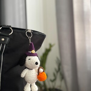 May include: A handmade crochet Snoopy keychain in a Halloween theme. The white Snoopy figure wears a purple witch hat and holds an orange trick-or-treat basket. The keychain is attached to a black tote bag.