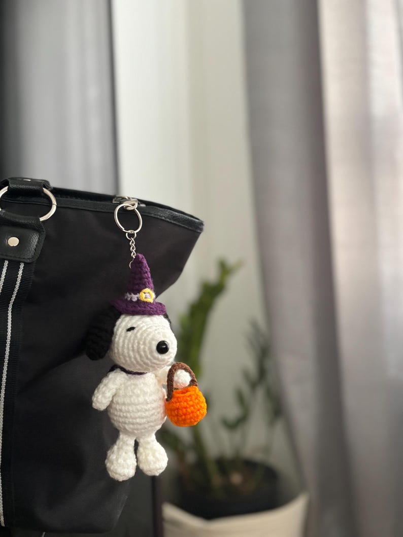 May include: A handmade crochet Snoopy keychain in a Halloween theme. The white Snoopy figure wears a purple witch hat and holds an orange trick-or-treat basket. The keychain is attached to a black tote bag.