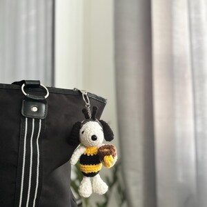May include: A handmade crochet keychain featuring a Snoopy-inspired character dressed as a bee. The figure has black and white details, yellow and black stripes, and holds a brown honey pot. The keychain is attached to a black tote bag.