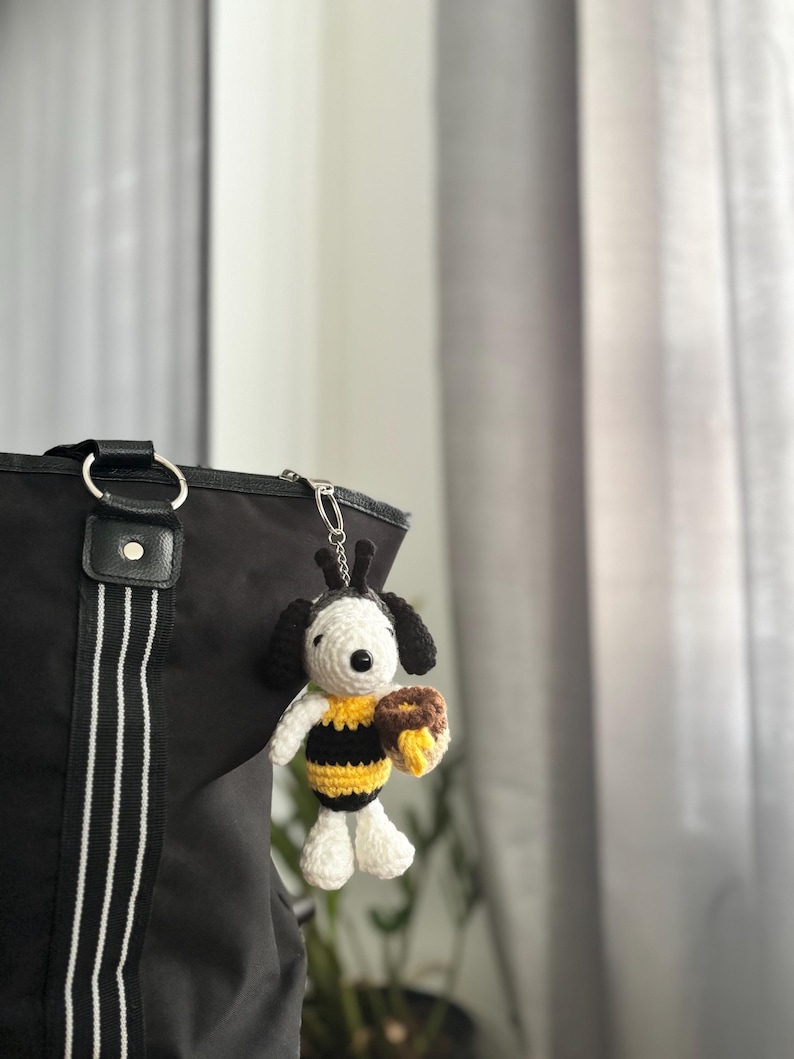 May include: A handmade crochet keychain featuring a Snoopy-inspired character dressed as a bee. The figure has black and white details, yellow and black stripes, and holds a brown honey pot. The keychain is attached to a black tote bag.