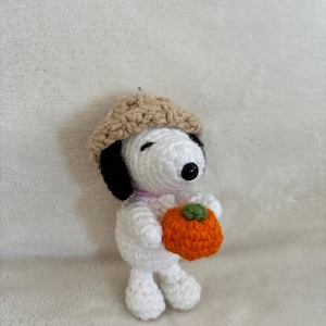 Op de afbeelding: Een handgemaakte gehaakte Snoopy-figuur met een beige baret en een oranje pompoen met een groene steel. De figuur is wit met zwarte oren en een zwarte neus. Een roze kraag zit om de nek.