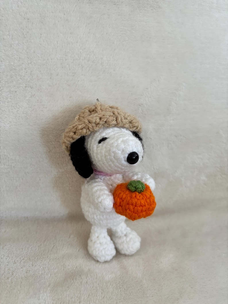 Op de afbeelding: Een handgemaakte gehaakte Snoopy-figuur met een beige baret en een oranje pompoen met een groene steel. De figuur is wit met zwarte oren en een zwarte neus. Een roze kraag zit om de nek.