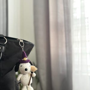 May include: A crocheted Snoopy keychain wearing a purple witch hat with a gold buckle and holding a small broom. The Snoopy figure is white with black ears and a black nose. The keychain is attached to a black bag.