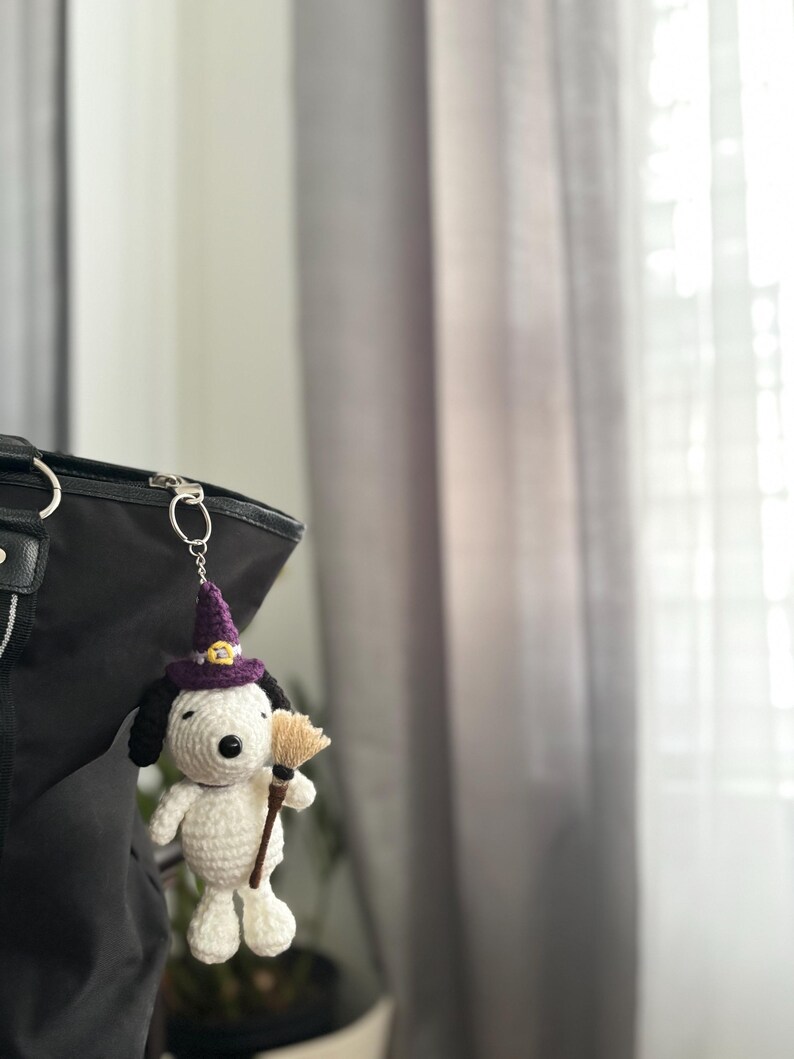 May include: A crocheted Snoopy keychain wearing a purple witch hat with a gold buckle and holding a small broom. The Snoopy figure is white with black ears and a black nose. The keychain is attached to a black bag.