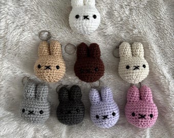 May include: A collection of eight crocheted rabbit keychains in various colors including white, tan, dark brown, cream, gray, charcoal, light purple, and pink. Each rabbit features two upright ears, two black bead eyes, and a stitched black X-shaped mouth.