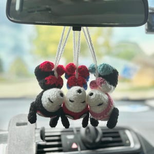 Op de afbeelding: Drie gehaakte pluche figuren hangend aan witte koorden. Het linkerfiguur is zwart en rood, het middelste figuur is rood en blauw en het rechterfiguur is zwart, wit en lichtblauw. Elk figuur heeft een wit gezicht met gesloten ogen.
