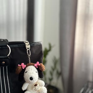 Op de afbeelding: Een handgemaakte gehaakte Snoopy sleutelhanger met bruine oren en een bruine bovenkant, versierd met roze strikjes. De witte Snoopy houdt een klein bruin voorwerp vast. De sleutelhanger is bevestigd aan een zwarte tas met een zilveren sluiting.