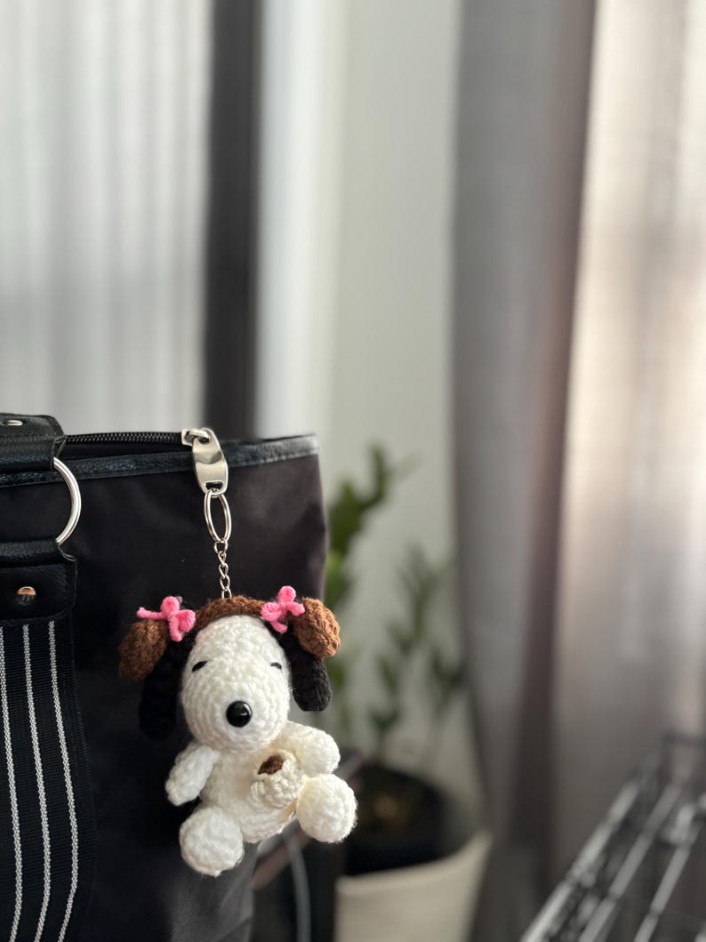 Op de afbeelding: Een handgemaakte gehaakte Snoopy sleutelhanger met bruine oren en een bruine bovenkant, versierd met roze strikjes. De witte Snoopy houdt een klein bruin voorwerp vast. De sleutelhanger is bevestigd aan een zwarte tas met een zilveren sluiting.