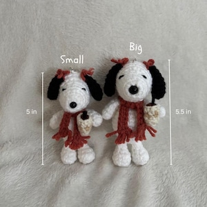 Op de afbeelding: Twee gehaakte Snoopy-poppen, &eacute;&eacute;n van 12,7 cm hoog en de andere van 14 cm hoog. Beide hebben zwarte oren, rode sjaals en houden een klein kopje vast. De poppen zijn wit met zwarte accenten en rode strikken.