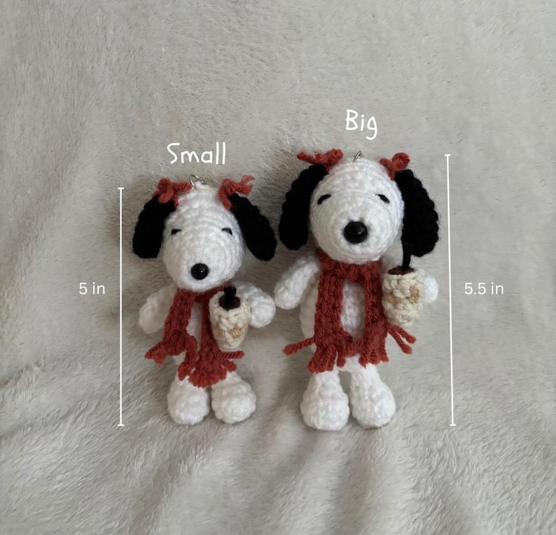 Op de afbeelding: Twee gehaakte Snoopy-poppen, &eacute;&eacute;n van 12,7 cm hoog en de andere van 14 cm hoog. Beide hebben zwarte oren, rode sjaals en houden een klein kopje vast. De poppen zijn wit met zwarte accenten en rode strikken.