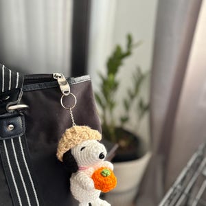 Op de afbeelding: Een handgemaakte gehaakte Snoopy sleutelhanger, met een witte body, zwarte oren en een beige baret. Snoopy houdt een oranje pompoen vast. De sleutelhanger is bevestigd aan een zwarte tas met een zilveren rits.