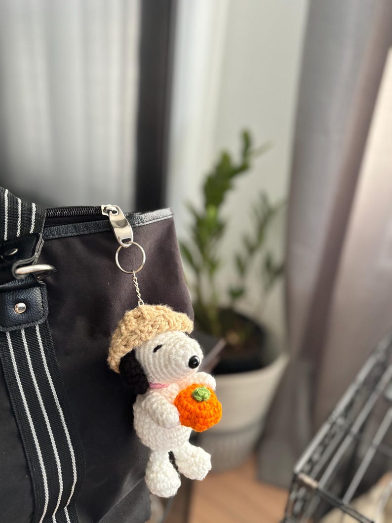 Op de afbeelding: Een handgemaakte gehaakte Snoopy sleutelhanger, met een witte body, zwarte oren en een beige baret. Snoopy houdt een oranje pompoen vast. De sleutelhanger is bevestigd aan een zwarte tas met een zilveren rits.