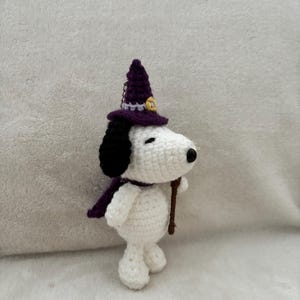 May include: A handmade crochet Snoopy figure wearing a purple witch hat and scarf, holding a brown stick. The figure is white with black ears and a black nose. The hat has a gold buckle. A Halloween-themed decorative item.