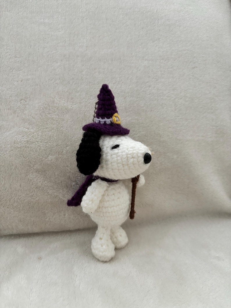 May include: A handmade crochet Snoopy figure wearing a purple witch hat and scarf, holding a brown stick. The figure is white with black ears and a black nose. The hat has a gold buckle. A Halloween-themed decorative item.