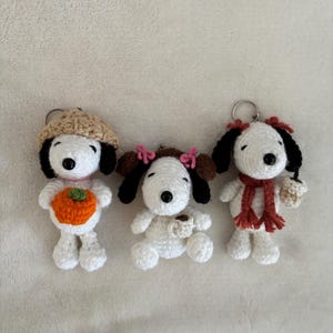 Op de afbeelding: Drie handgemaakte Snoopy-sleutelhangers van gehaakt garen. E&eacute;n sleutelhanger draagt een beige hoed en houdt een oranje pompoen vast. Een andere sleutelhanger heeft roze strikjes en een koffiekopje. De derde sleutelhanger heeft een rode sjaal en een koffiekopje.